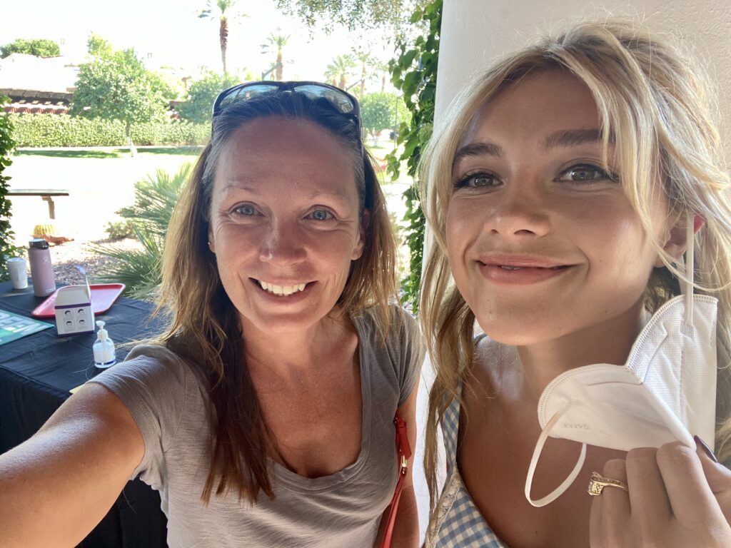 Two women smile for a selfie. The one on the right has a mask on her ear.