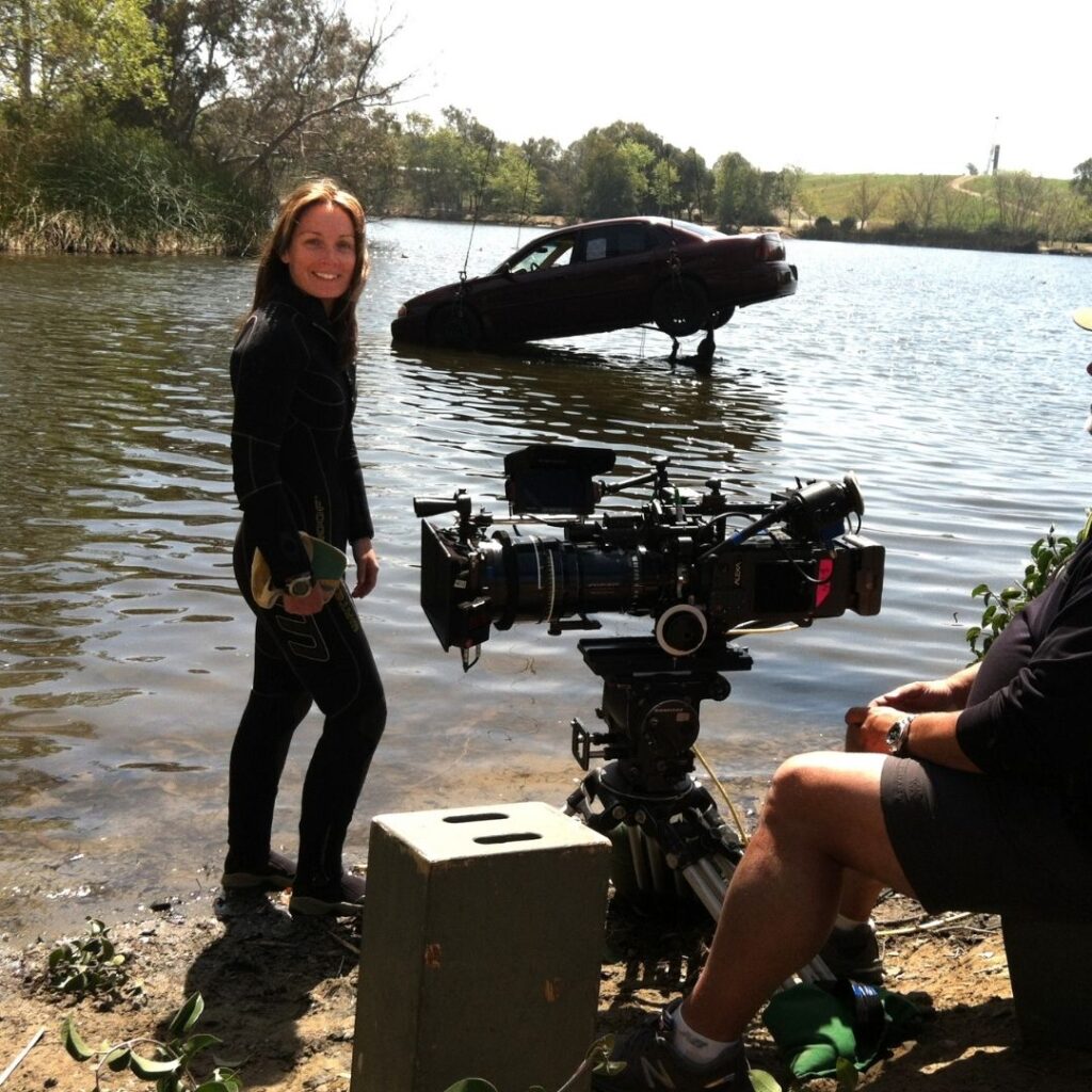 A female stuntwoman stands at the water's edge, with a prop car about to hit the water. A camera and cameraman are in the foreground.