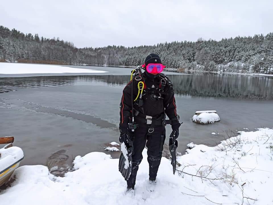 A female scuba diver stands in the snow on the side of a Finnish lake