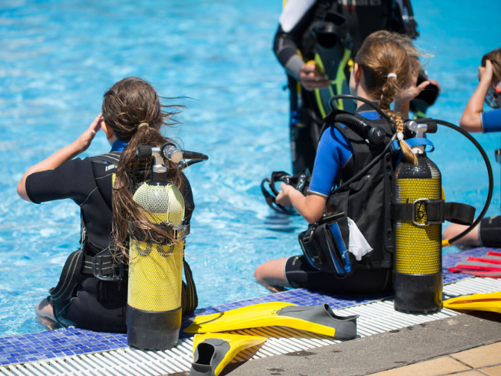 two kids prepare to go scuba diving in a swimming pool. They are sitting on the edge of the pool facing away from the camera.