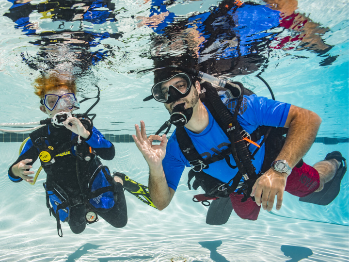 a young scuba diver and his instructor are in a swimming pool and giving the okay sign towards the camera