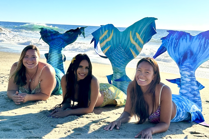 PADI Professional Mermaids Elle Jimenez, Elaina Thomas, and Great Chin Burger posing on a sandy beach next to the ocean