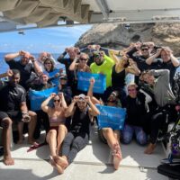 The PADI AWARE team sit and stand on a boat and hold a PADI AWARE sign while giving the scuba diver signal for shark, which is a hand with fingers held together vertically with the thumb side of the hand placed on the forehead between the eyes