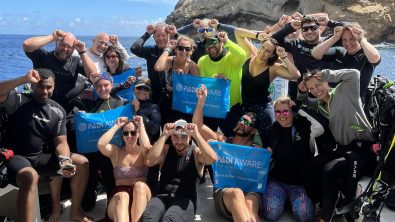 The PADI AWARE team sit and stand on a boat and hold a PADI AWARE sign while giving the scuba diver signal for shark, which is a hand with fingers held together vertically with the thumb side of the hand placed on the forehead between the eyes