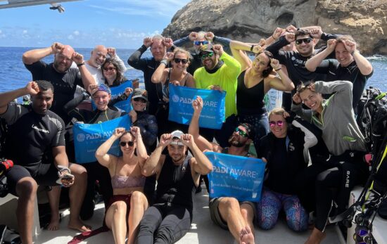 The PADI AWARE team sit and stand on a boat and hold a PADI AWARE sign while giving the scuba diver signal for shark, which is a hand with fingers held together vertically with the thumb side of the hand placed on the forehead between the eyes