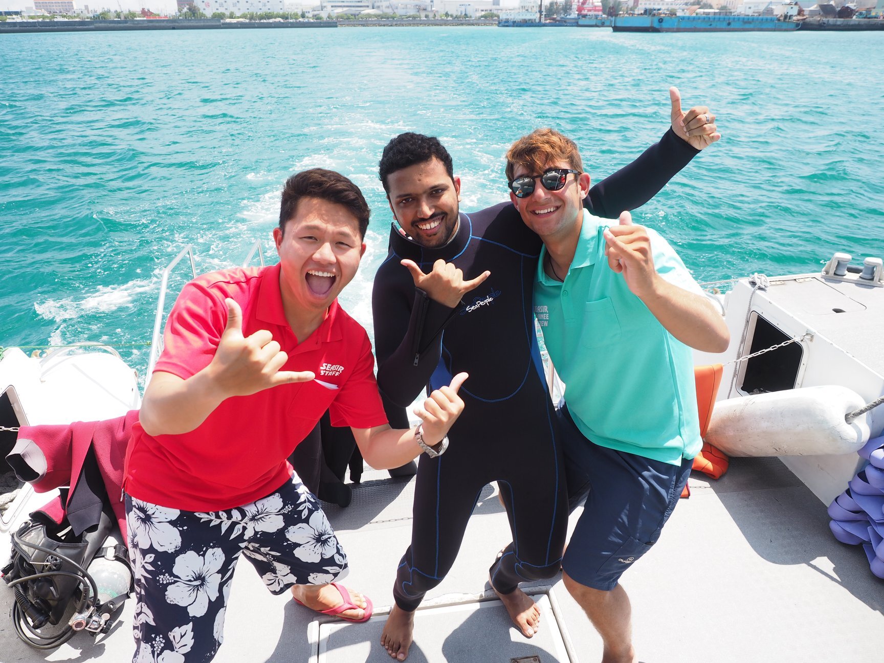 three divers are standing on a boat in the tropics giving the hang loose sign. They look very happy and excited.