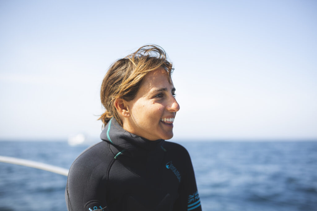 A woman in a black wetsuit smiles with the ocean in the background.