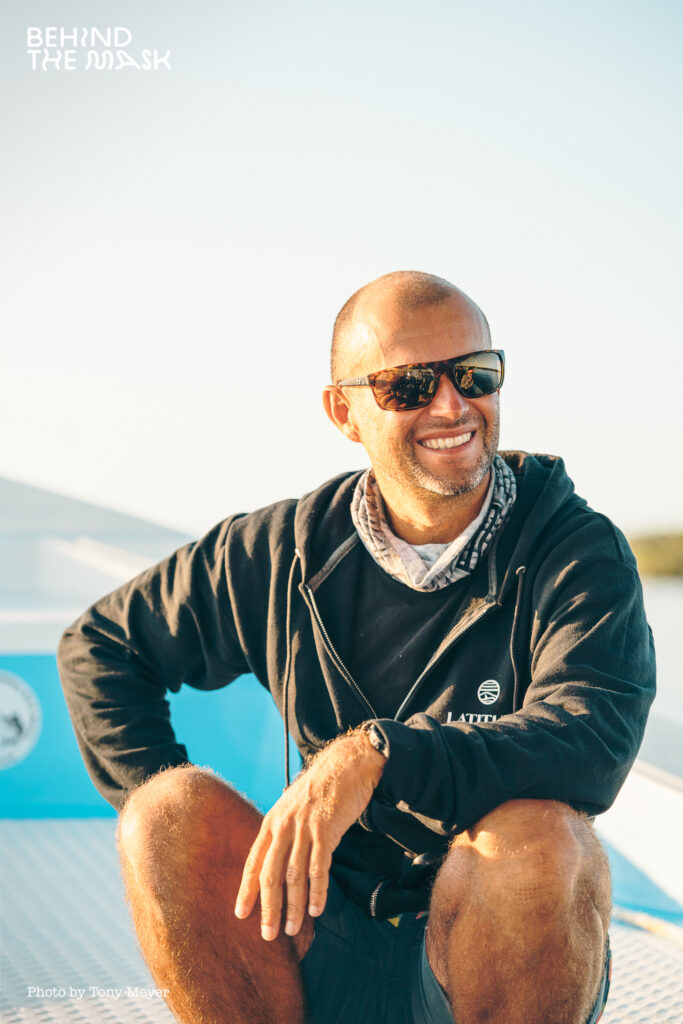 A man in sunglasses and a long-sleeved shirt smiles in the sunshine on a boat.