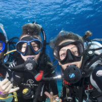 three divers are underwater taking a selfie together. Two of the divers give the hang loose symbol with their right hands.