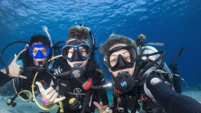 three divers are underwater taking a selfie together. Two of the divers give the hang loose symbol with their right hands.