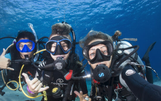 three divers are underwater taking a selfie together. Two of the divers give the hang loose symbol with their right hands.