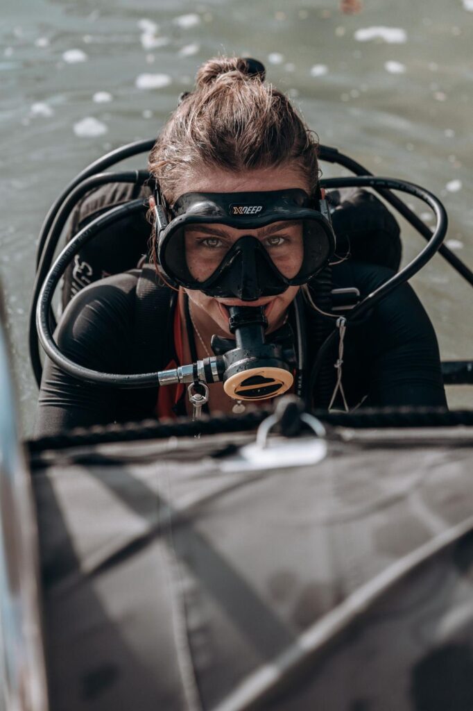 A woman diver emerges from the water next to a boat. She has her gear still on, including her regulator in her mouth.