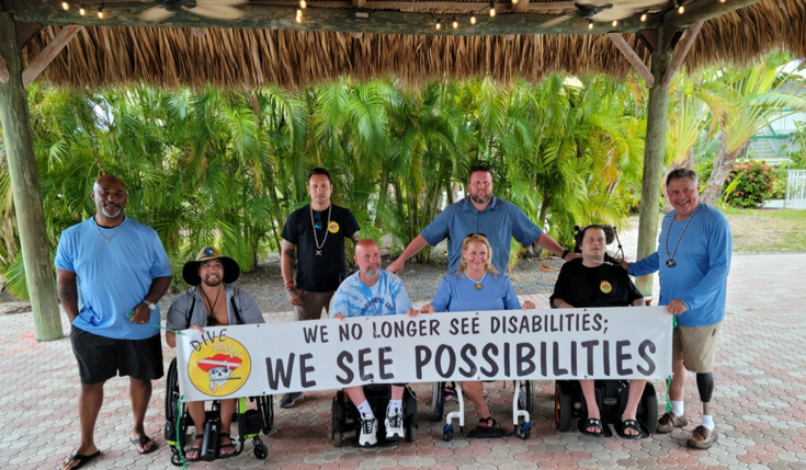 A group of differently abled divers pose together holding a sign that says, "We no longer see disabilities; we see possibilities."