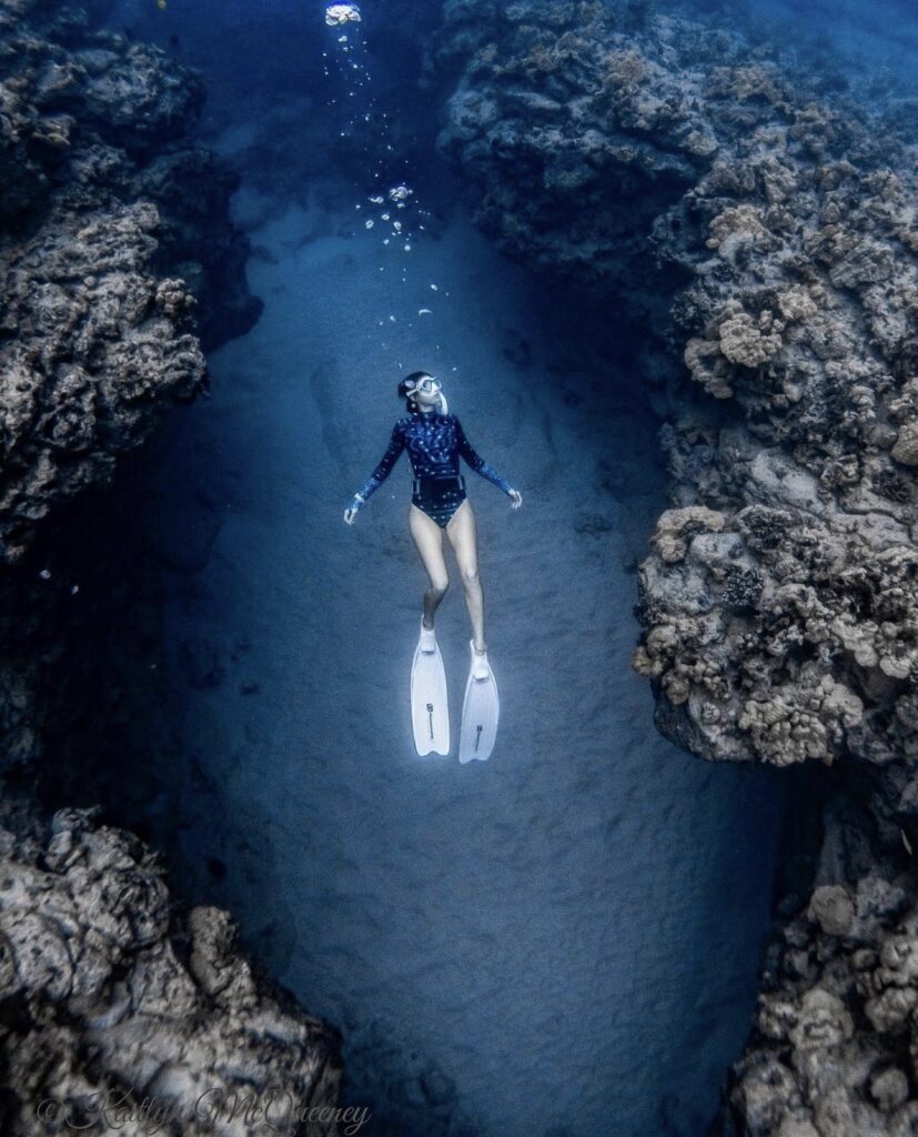 A female freediver floats above the sand, between two coral mounds. She wears a whalesuit bodysuit and white fins.