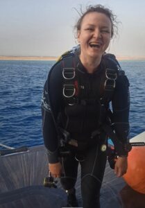 A woman stands smiling on a boat in dive gear