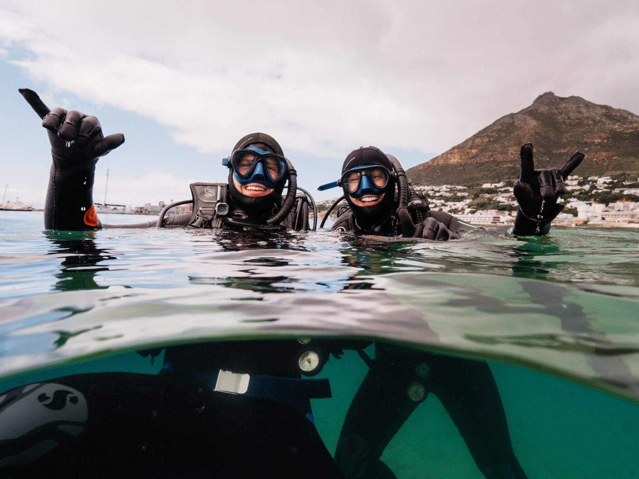 Two scuba divers are at the surface of the water with Cape Town in the background. They are holding up hang loose signs with their hands.