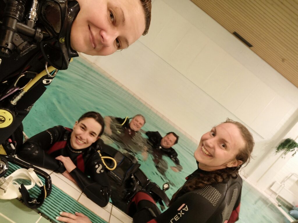 Three female divers sit at the side of the pool and smile of the camera, selfie style