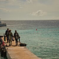 three scuba divers walk to the end of a dock. there is a boating anchored offshore.
