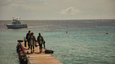 three scuba divers walk to the end of a dock. there is a boating anchored offshore.