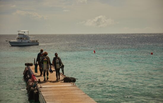 three scuba divers walk to the end of a dock. there is a boating anchored offshore.