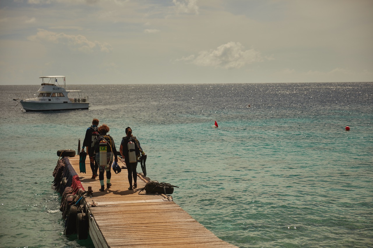 three scuba divers walk to the end of a dock. there is a boating anchored offshore. 