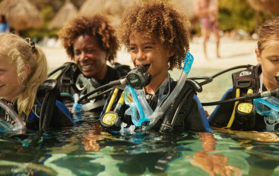 a young scuba diver stands in waist deep water in scuba gear with other kids and an instructor looking on