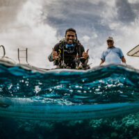 A scuba instructor sits on a boat and gives two shakas to the photographer in the water