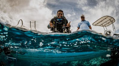 A scuba instructor sits on a boat and gives two shakas to the photographer in the water