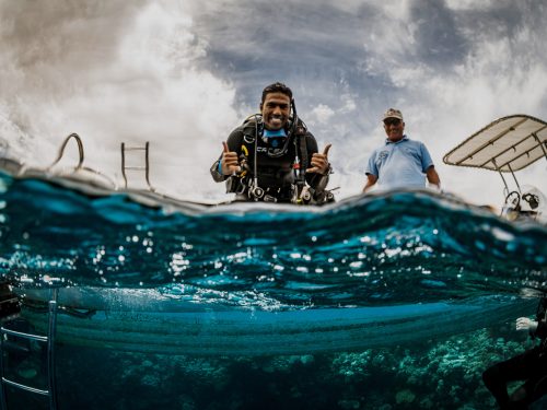 A scuba instructor sits on a boat and gives two shakas to the photographer in the water