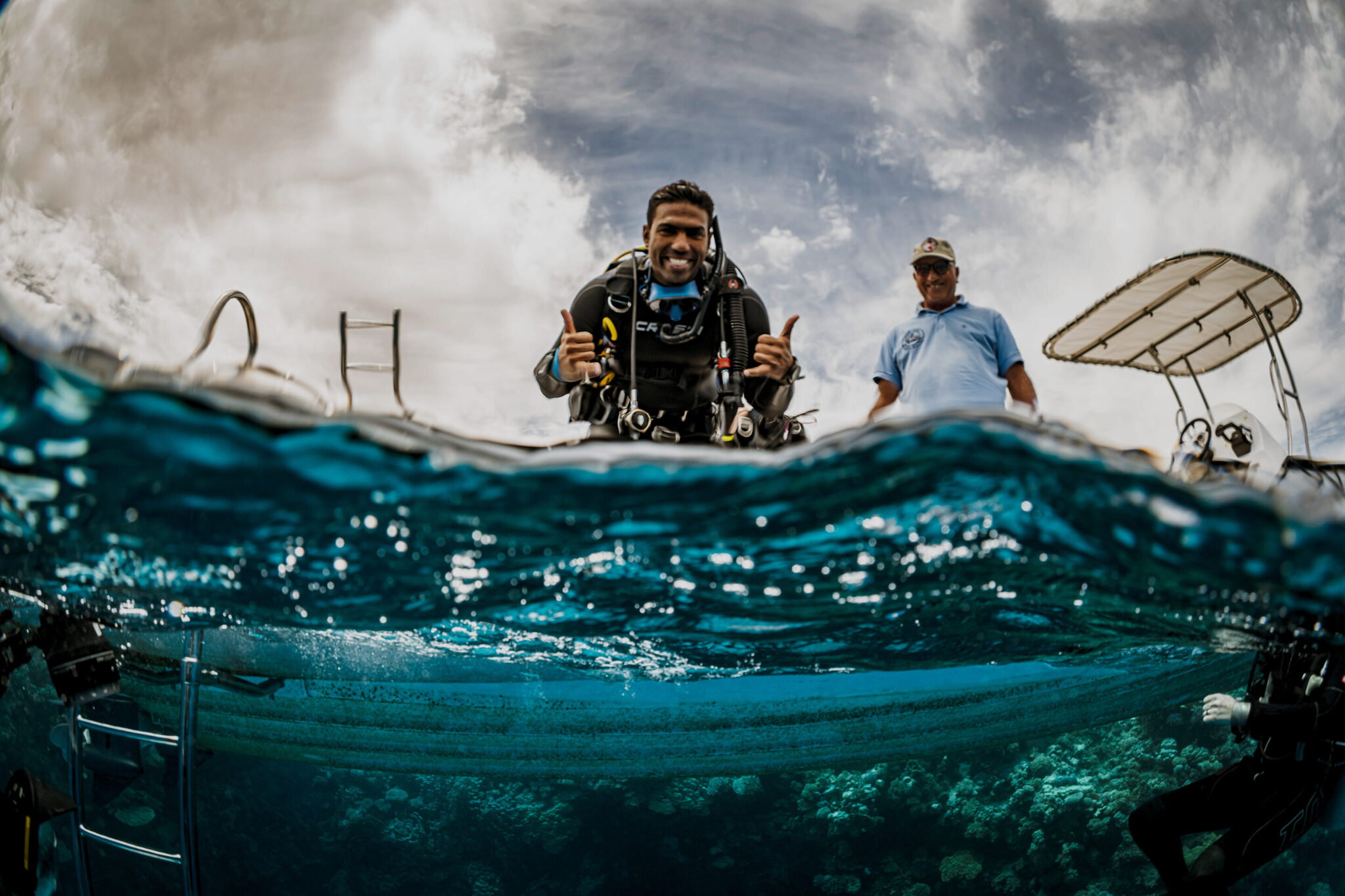 A scuba instructor sits on a boat and gives two shakas to the photographer in the water