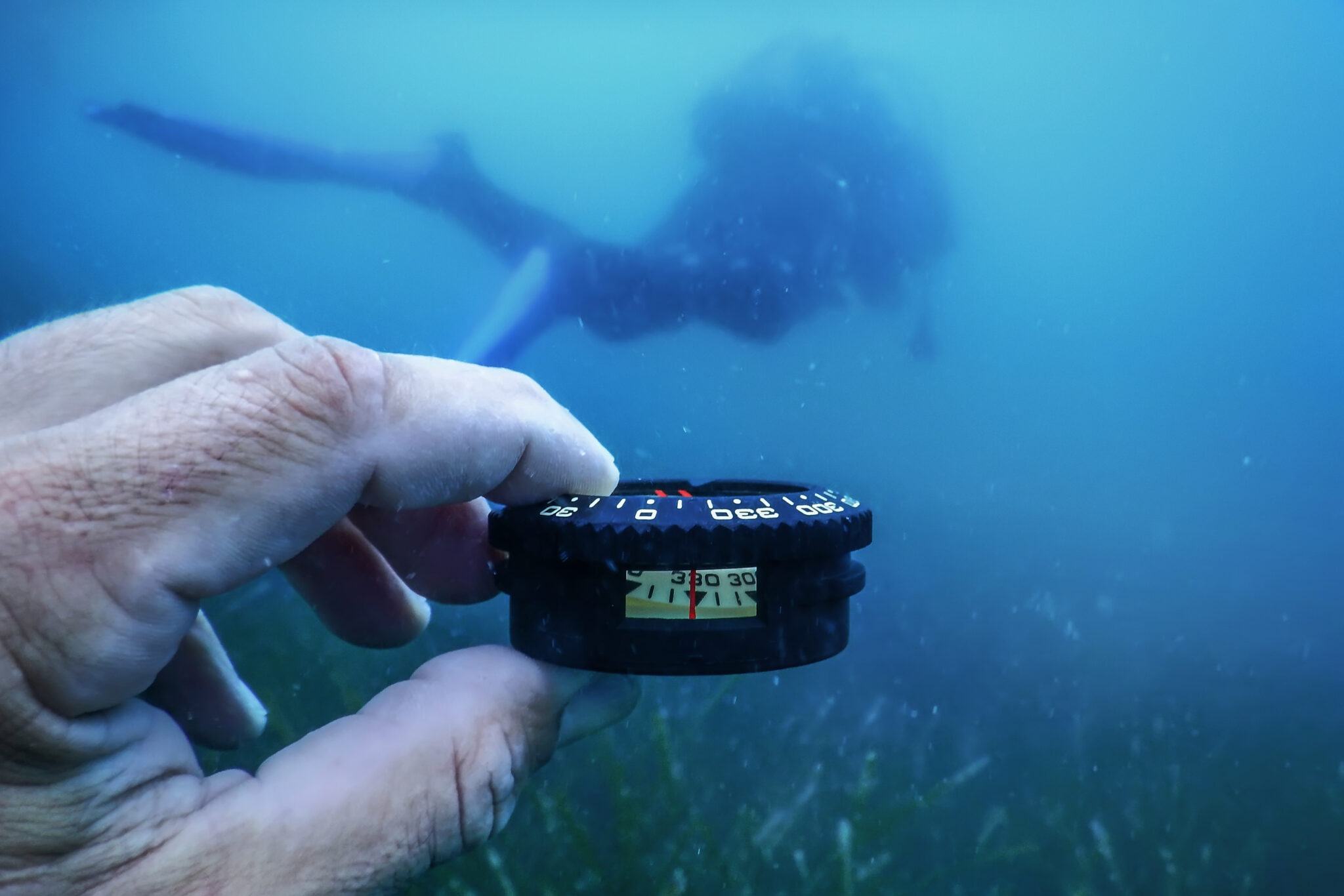a scuba diver looks at his underwater compass to aid in underwater navigation. Another diver is in front of the compass.
