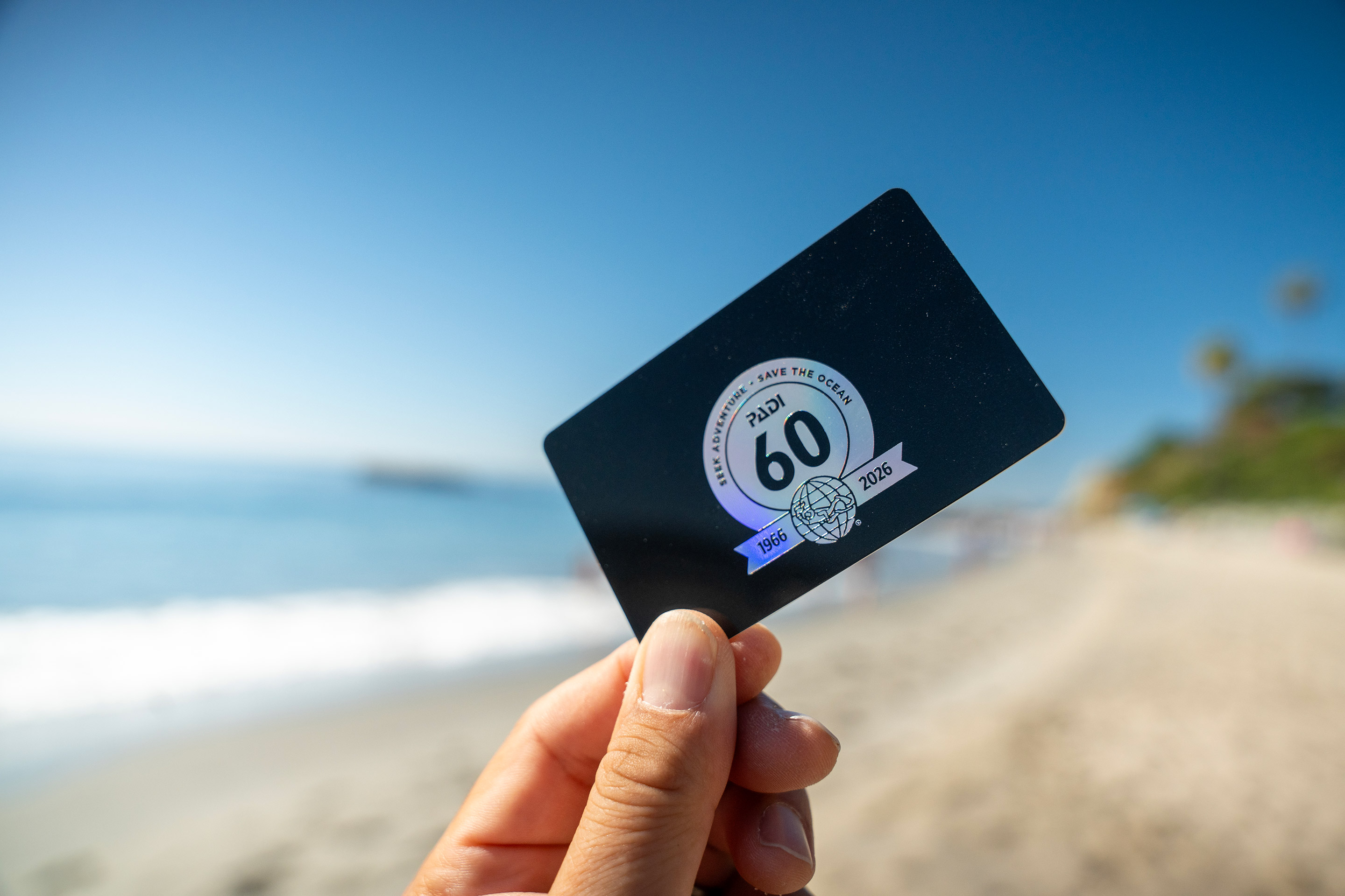 A hand holds up a black 60th anniversary certification card on a beach
