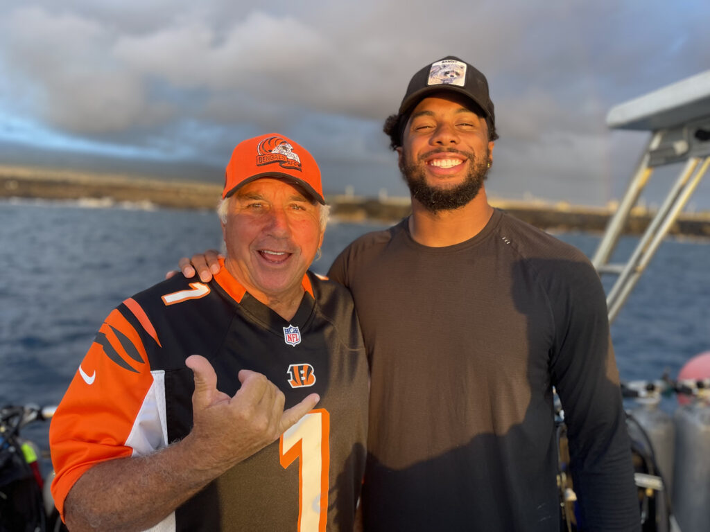 Two men smile on a dive boat. One has an NFL jersey for the Cincinnati Bengals and the one on the right is a player on the team.