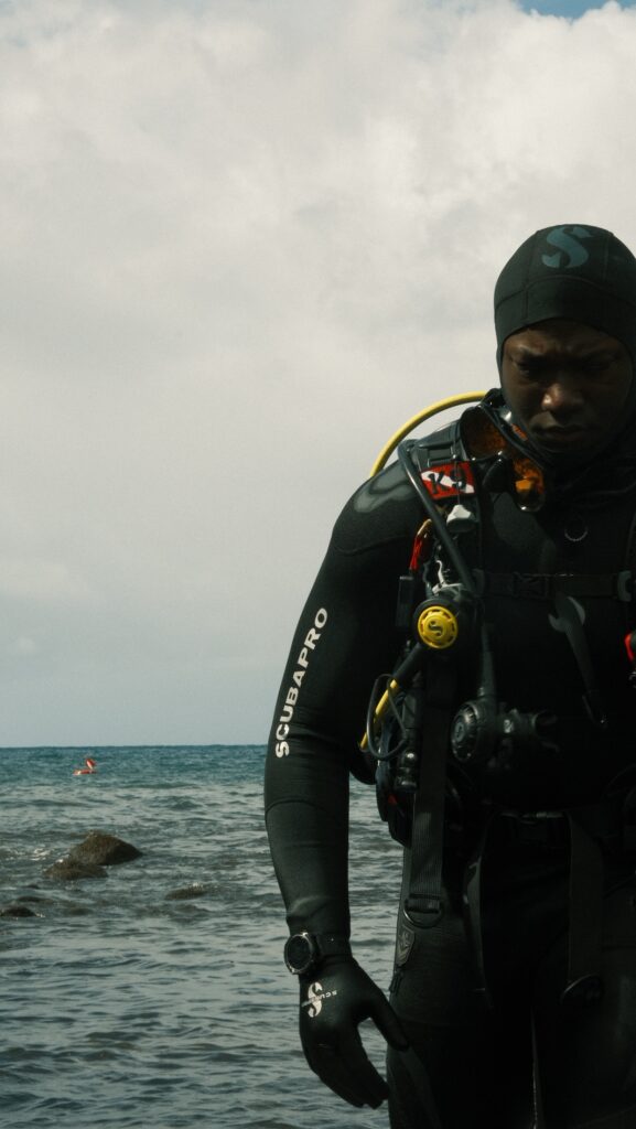 A diver walks out of the ocean after a shore dive looking down. He is wearing all black.