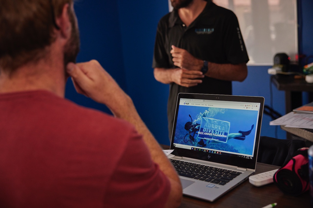 a man in a dive shop looks at a laptop showing the PADI AWARE Foundation homepage