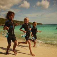 three kids run along the beach. they are wearing wetsuits and carrying masks and snorkels
