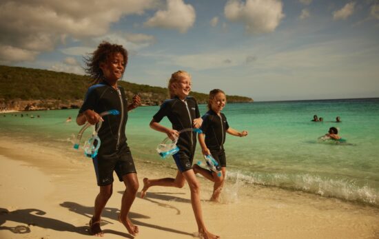 three kids run along the beach. they are wearing wetsuits and carrying masks and snorkels