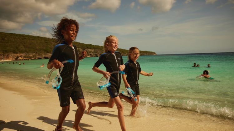 three kids run along the beach. they are wearing wetsuits and carrying masks and snorkels