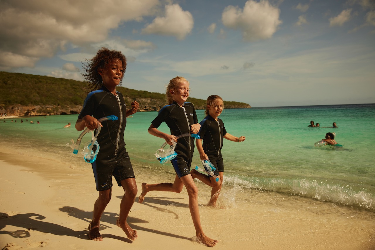 three kids run along the beach. they are wearing wetsuits and carrying masks and snorkels