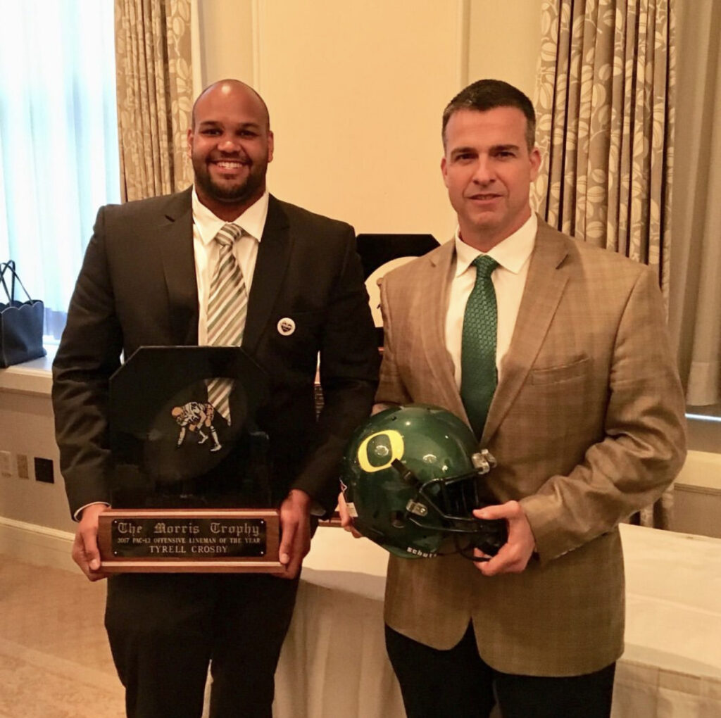 Two men stand holding (1) an award and (2) an Oregon State Football helmet