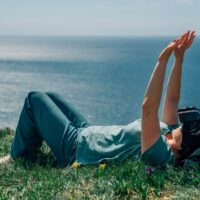 A person lying on their back on a grassy hill overlooking the ocean. They wear VR goggles and have their arms outstretched