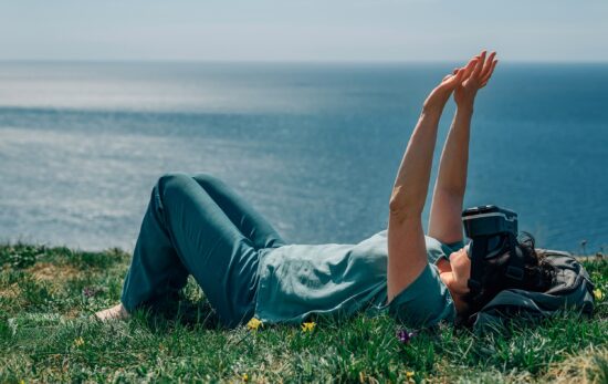 A person lying on their back on a grassy hill overlooking the ocean. They wear VR goggles and have their arms outstretched