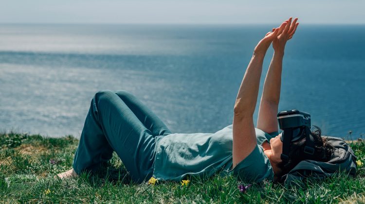 A person lying on their back on a grassy hill overlooking the ocean. They wear VR goggles and have their arms outstretched
