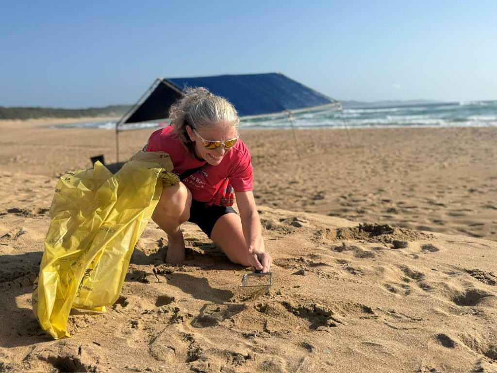 Amanda van Jaarsvled kneeling on a beach sifting through sand to clean up plastic