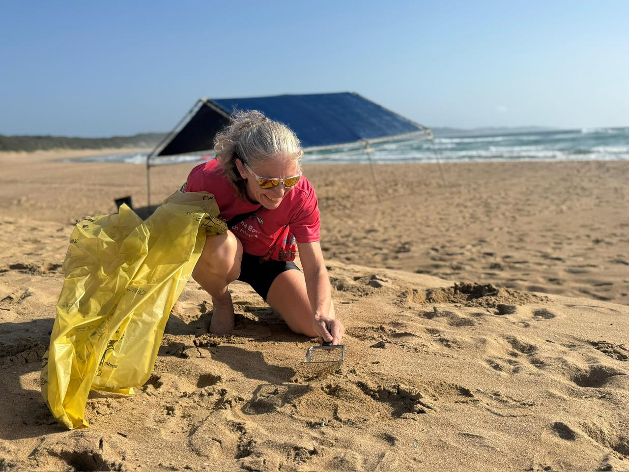 Amanda van Jaarsvled kneeling on a beach sifting through sand to clean up plastic
