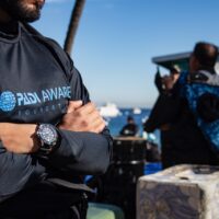 a man stands on a beach with his arms crossed. he is wearing a padi aware rashguard and a seiko watch