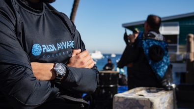 a man stands on a beach with his arms crossed. he is wearing a padi aware rashguard and a seiko watch