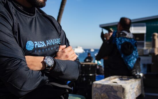 a man stands on a beach with his arms crossed. he is wearing a padi aware rashguard and a seiko watch