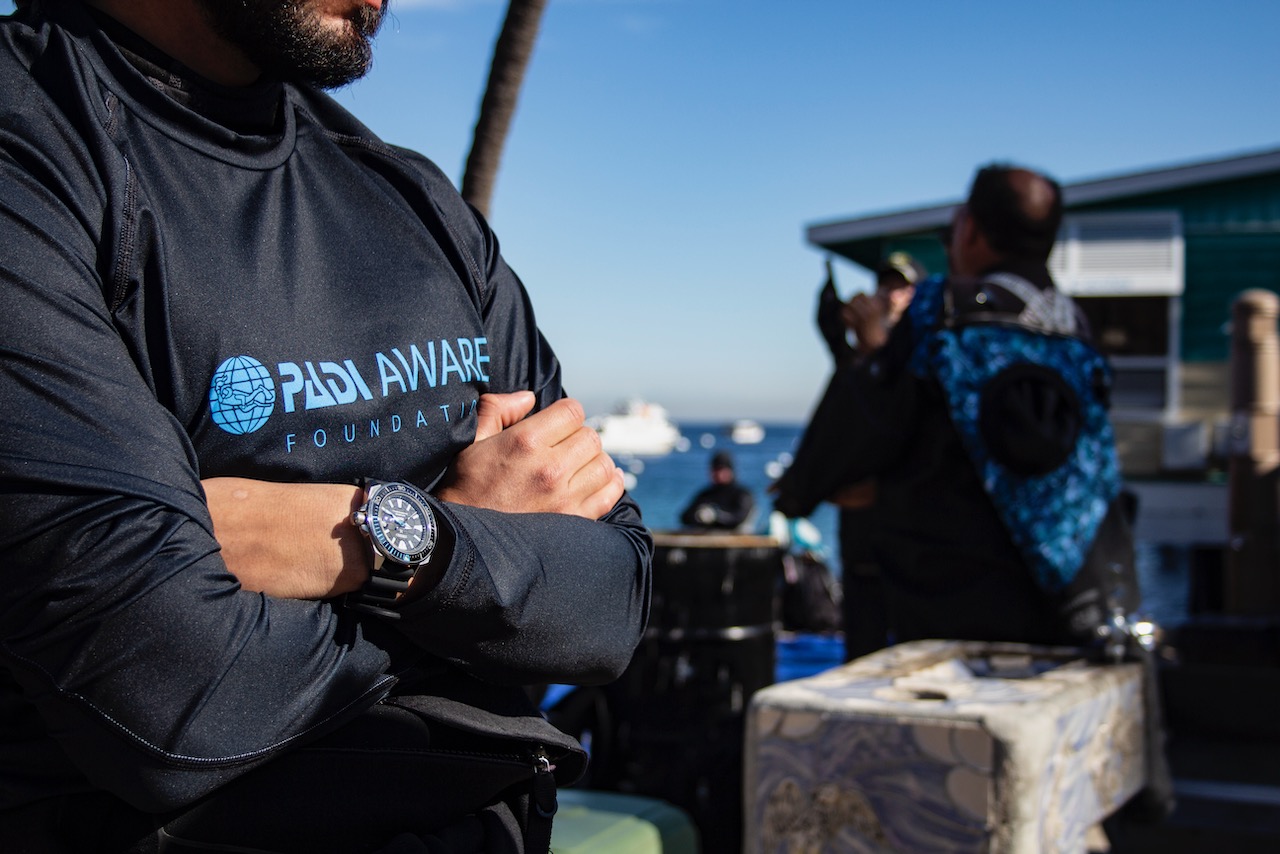 a man stands on a beach with his arms crossed. he is wearing a padi aware rashguard and a seiko watch