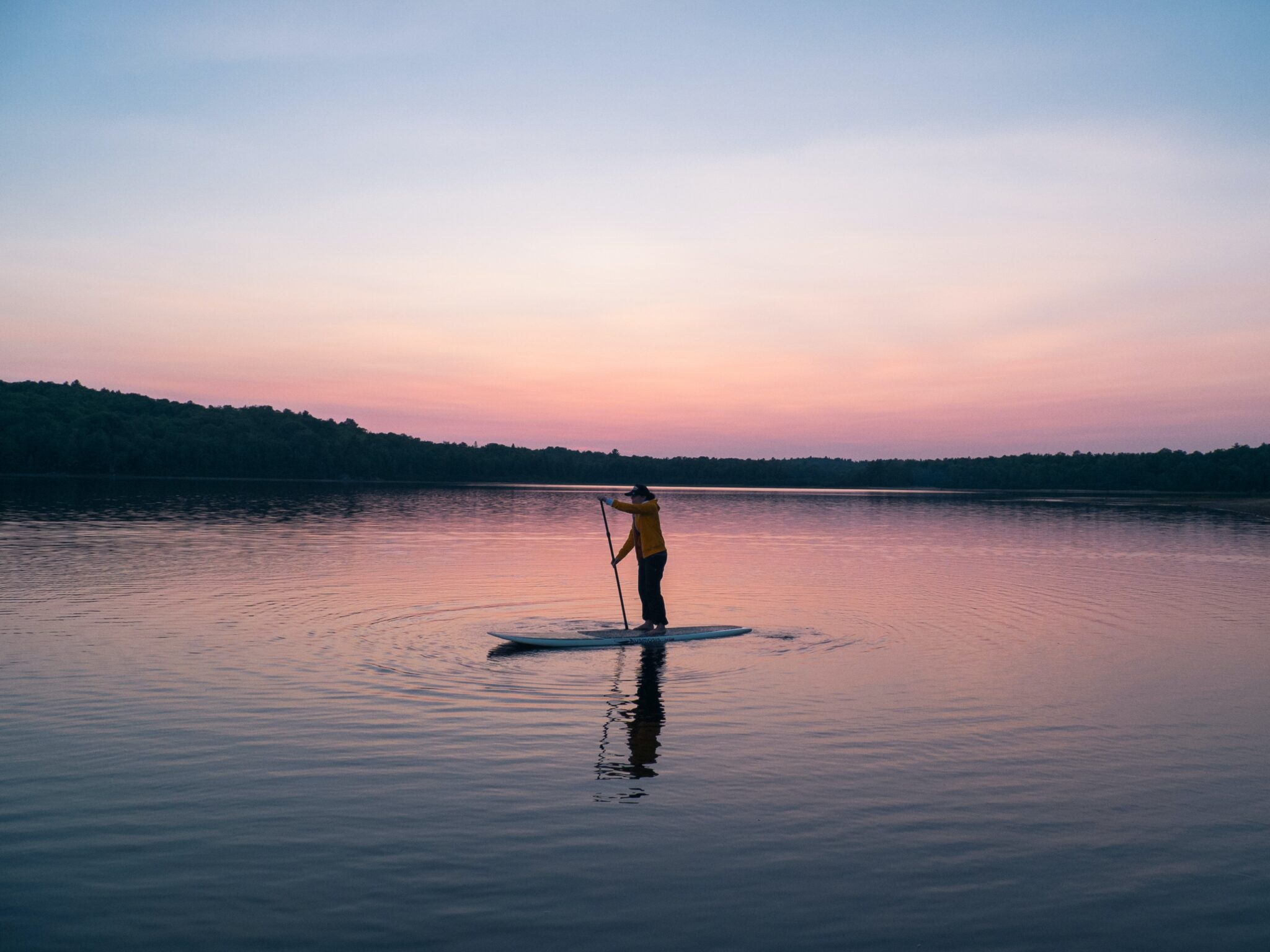 A woman uses a paddleboard on a lake at sunset. She is wearing a jacket and heavy pants.